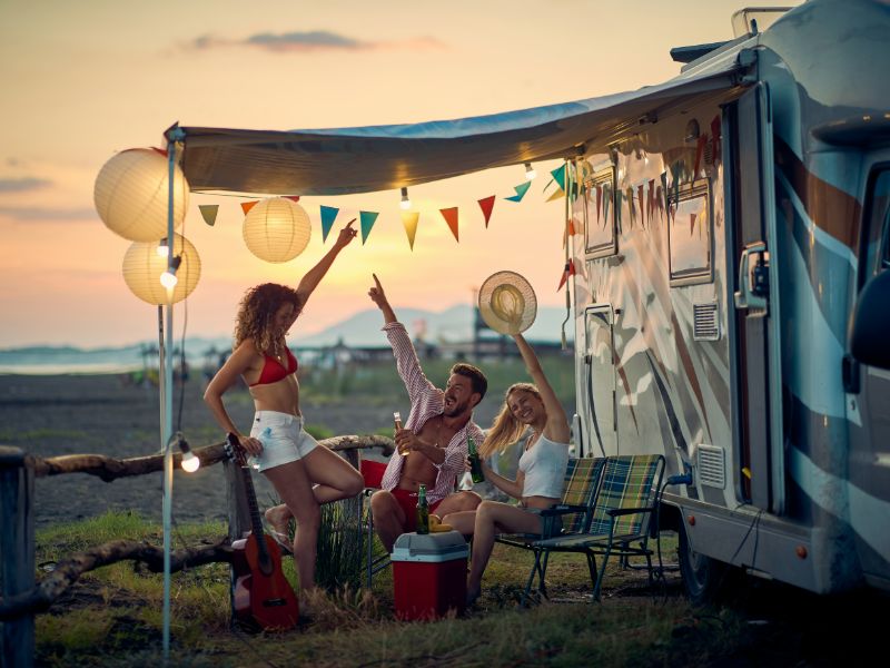 Drei Freunde vor mit Fahnen verziertem Camper mit alkoholischen Getränken und Gitarre.