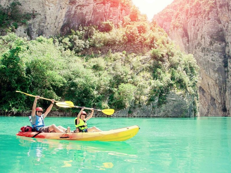 Mann und Kind in Kanu mit vor Freude gestreckten Armen mit Kanu-Paddel in den Händel auf türkisblauem Wasser mit Felsen im Hintergrund.